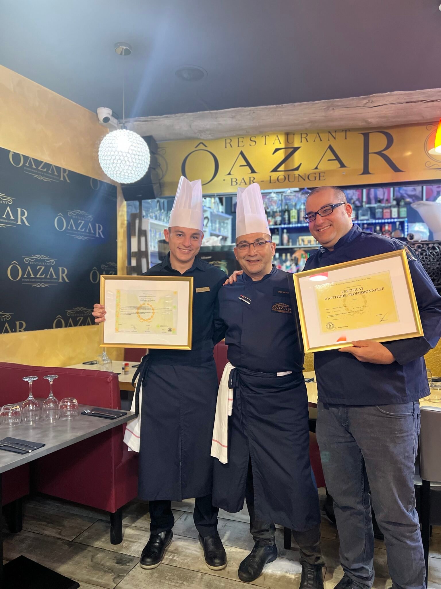 Trois directeurs père et fils en tenue posent fièrement dans le restaurant Ôazar à Cavaillon, tenant leurs diplômes culinaires.